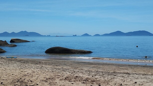 A beach with large rocks, calm water, and distant mountains.