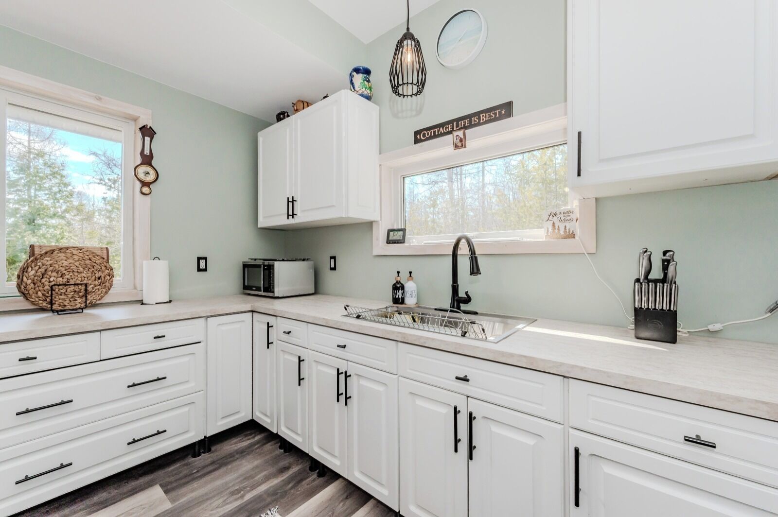 A modern kitchen with white cabinets, a microwave, a sink, and a window with a view of trees.
