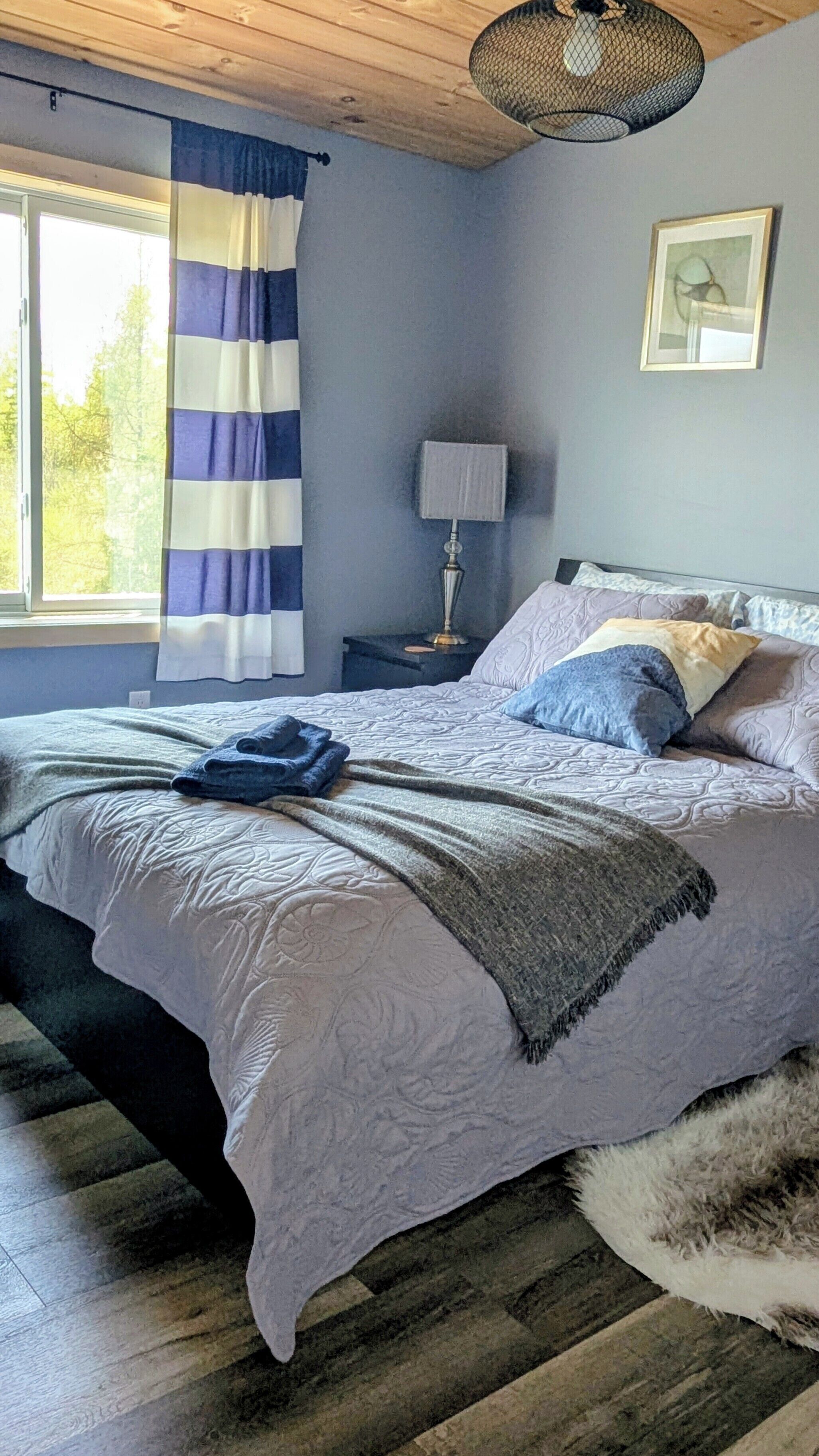 A bedroom with a bed, a nightstand, a lamp, a window with curtains, and a picture on the wall.