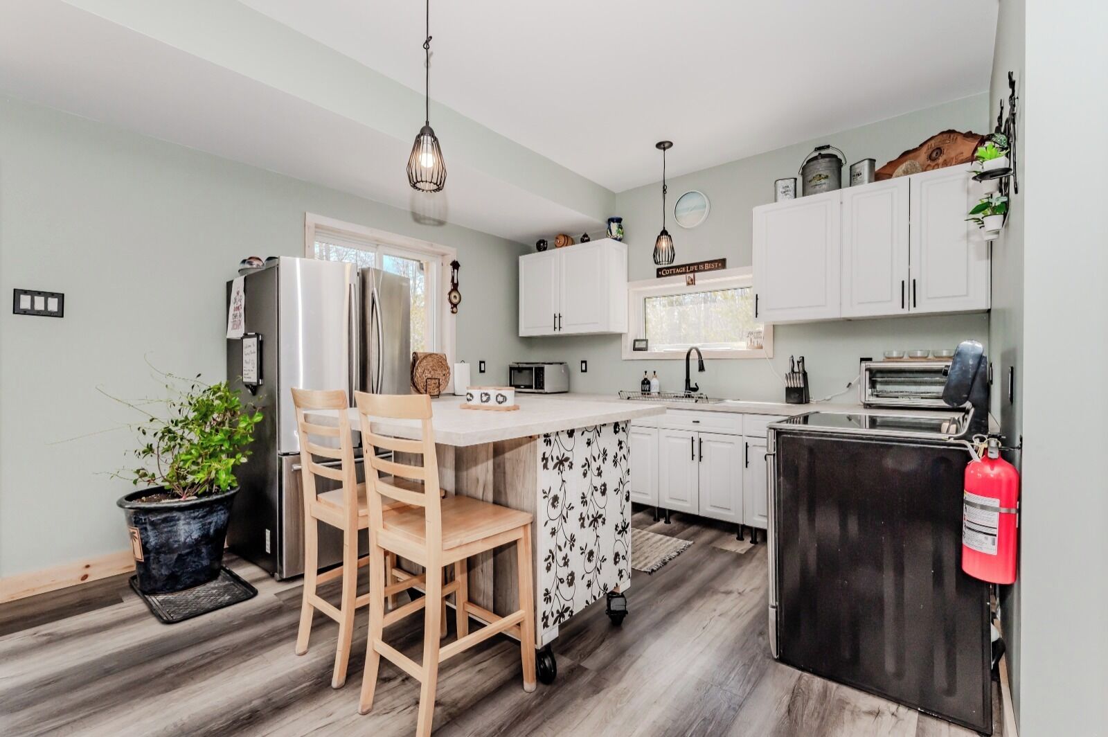 A compact kitchen with white cabinets, a central island, wooden chairs, a potted plant, and a fire extinguisher.