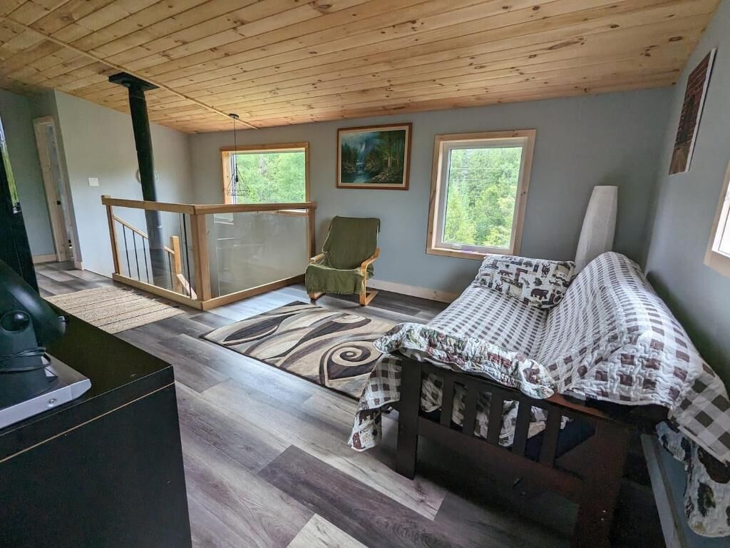A cozy cabin interior with a bed, a chair, a fireplace, and a view of the outdoors through large windows.