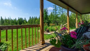 A wooden deck with a railing, surrounded by potted plants and flowers, overlooking a grassy area with trees.