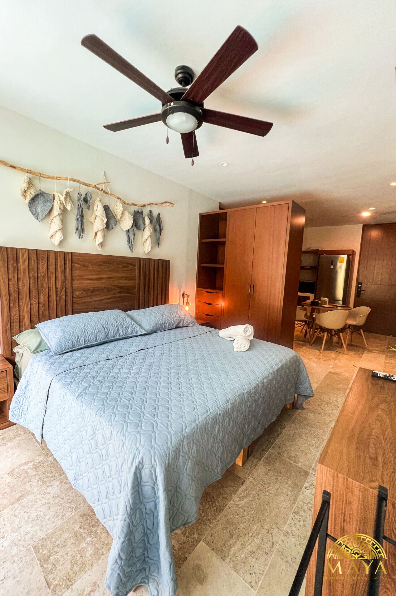 Un dormitorio con cama, ventilador de techo, cabecero de madera, estante en la pared y escritorio con sillas.
