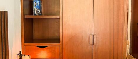 Un dormitorio con una cama, un armario de madera con varias repisas, una lámpara de noche y una repisa en la pared con un libro.