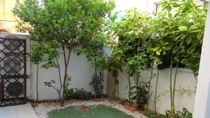 Un pequeño patio con césped artificial verde, rodeado de diversas plantas y árboles, y un muro blanco con una reja de metal.