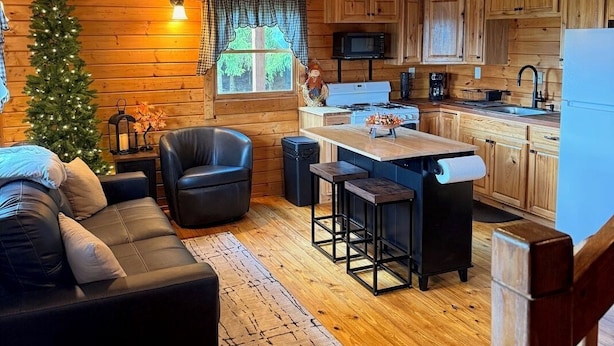 L’intérieur chaleureux d’un chalet avec un plafond en bois, un canapé en cuir noir, un petit sapin de Noël, un coin cuisine doté de meubles en bois et d’un four à micro-ondes, et un ventilateur de plafond.