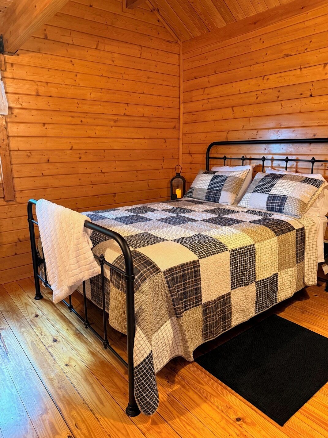 Une chambre dans une cabane en bois, avec un lit en métal, une couvre-lit matelassée et un tapis noir.