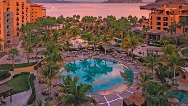 Een resort met een centraal gelegen zwembad, palmbomen en uitzicht op zee bij zonsondergang.