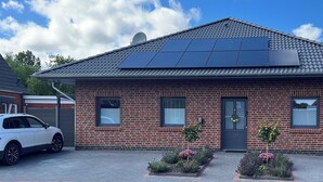 Une maison en briques avec un toit équipé de panneaux solaires, une voiture garée devant et un petit jardin fleuri.