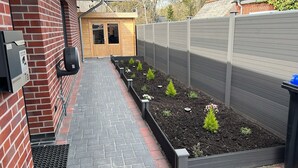 Un étroit sentier menant à une porte en bois, bordé d’un mur de briques et d’une clôture en métal.