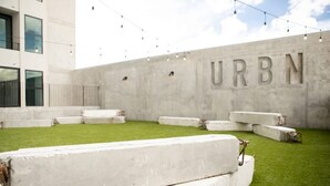 Um pátio com um muro de concreto identificado com a inscrição 'URBN' e bancos brancos dispostos em semicírculo.