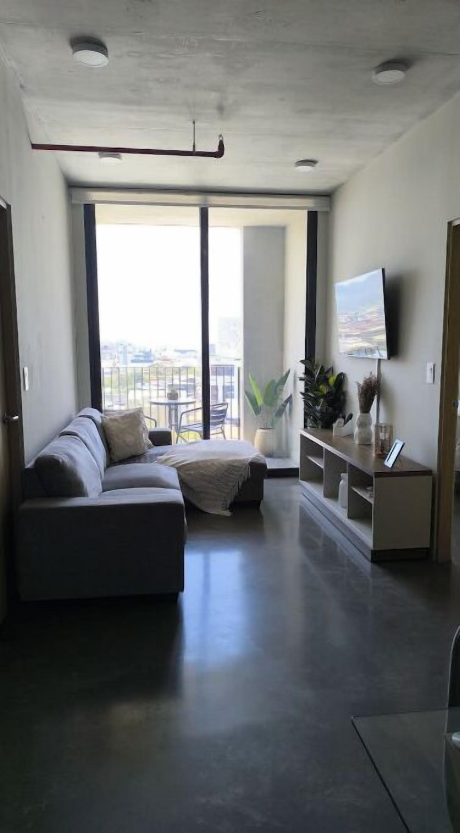 Una sala de estar moderna con un sofá seccional, un televisor de pantalla plana montado en la pared y un balcón con vistas.