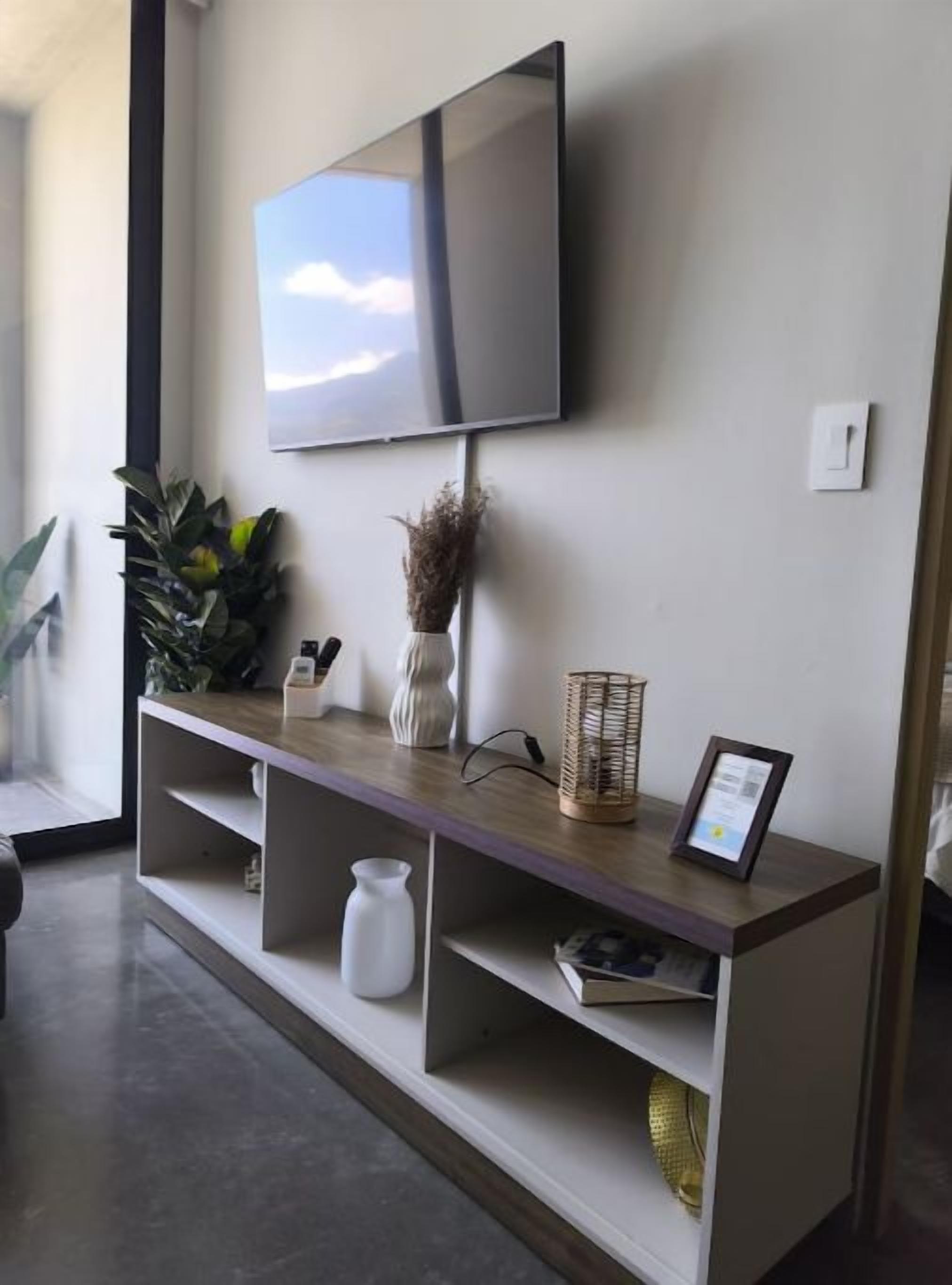 A modern living room with a flat-screen TV mounted on the wall, a wooden console table with decorative items, and a large window providing natural light.