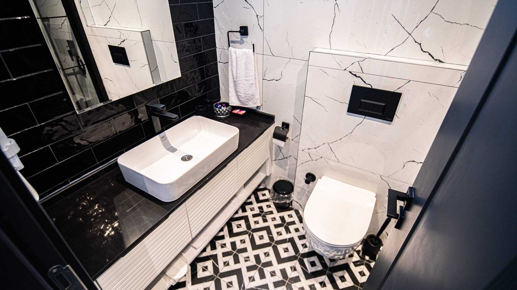 Un baño moderno con un lavamanos blanco, encimera negra y un inodoro. El piso presenta un estampado geométrico en blanco y negro.