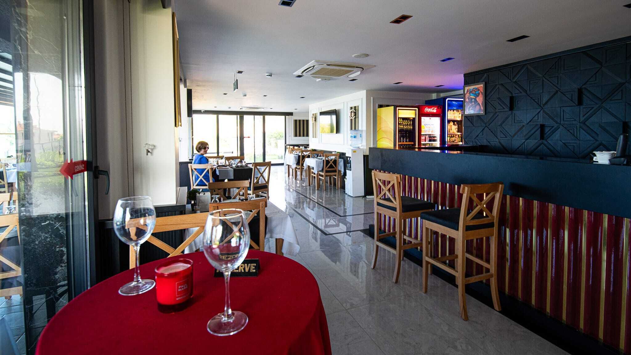 Un ristorante con zona bar, sedie e tavoli in legno. Si vedono bicchieri da vino su un tavolo, e una persona sullo sfondo.