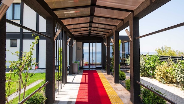 Un pasillo cubierto con una alfombra roja que conduce a una puerta de cristal, rodeado de follaje y vista al mar.