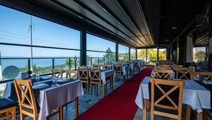 Un restaurante con mesas preparadas para comer, amplias ventanas que dan a una vista del mar, y una alfombra roja que conduce al área de asientos.
