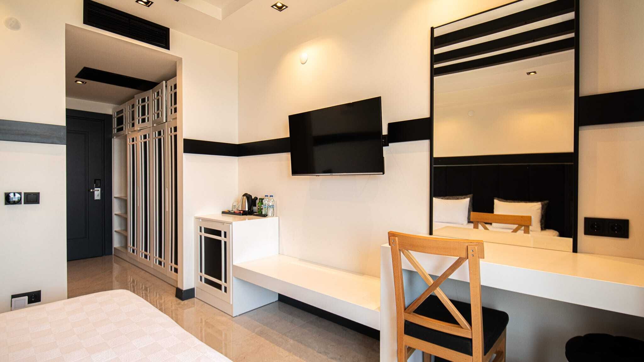 Una habitación de hotel moderna con una cama grande, un televisor montado en la pared, una silla de madera y una mesita con una tetera y botellas.