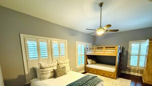Une chambre avec un lit, un lit superposé, un ventilateur de plafond et des fenêtres équipées de stores.