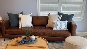 Un canapé en cuir marron agrémenté de plusieurs coussins, une table basse en bois ornée d’objets décoratifs et deux poufs posés sur un tapis à motifs.