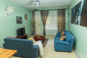 Ein Wohnzimmer mit zwei blauen Sofas, einem Holztisch, einem Fernseher, einem Ventilator und einem Bild an der Wand.