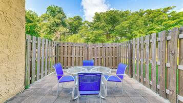 Une terrasse avec une table ronde en verre et quatre chaises bleues, entourée d’une clôture en bois et d’un mur de couleur beige.