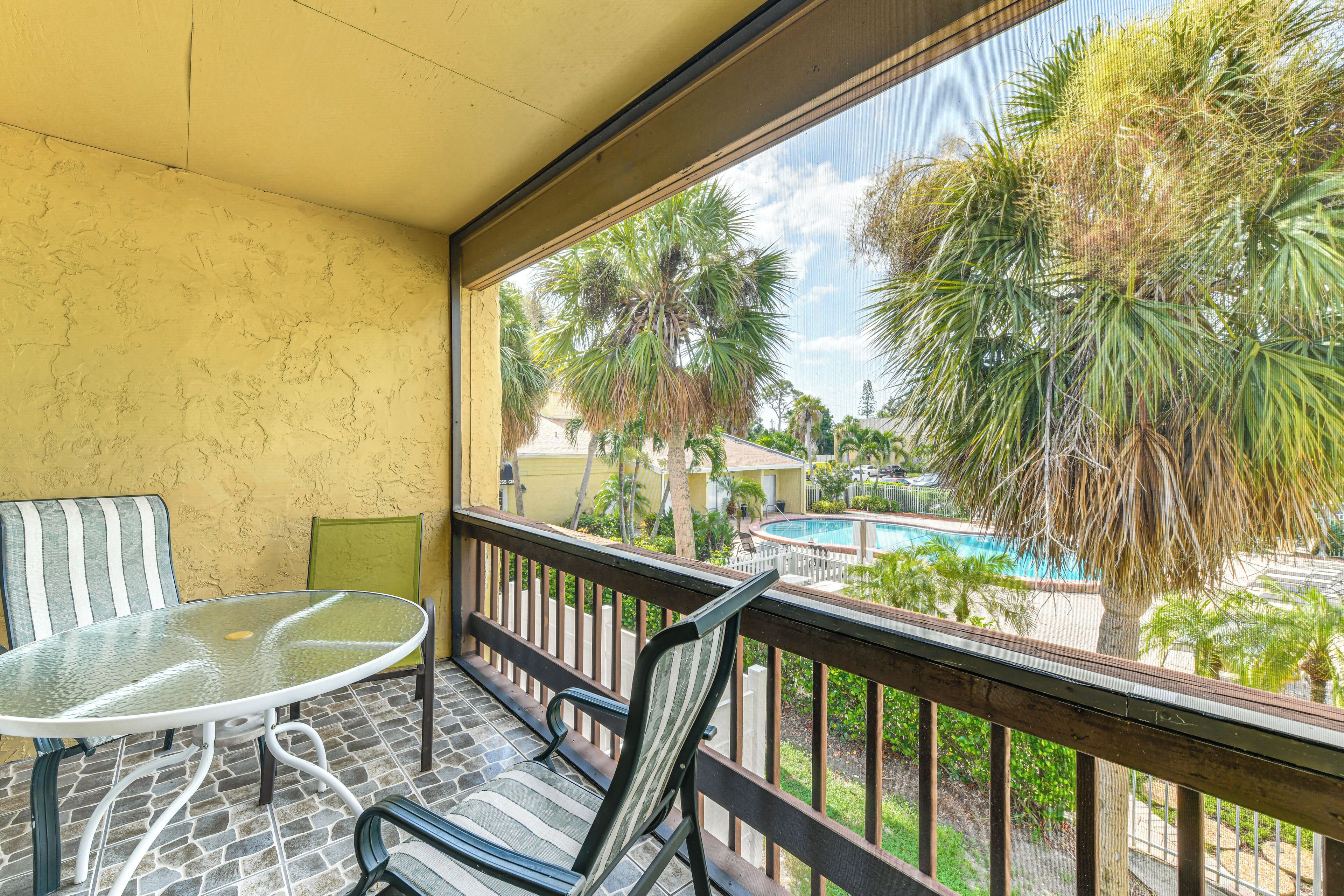 A balcony with a glass table and chairs, overlooking a swimming pool and palm trees.