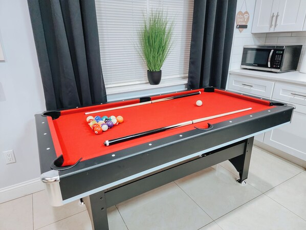 A pool table with balls and cues, a potted plant, and a microwave in a modern kitchen area.