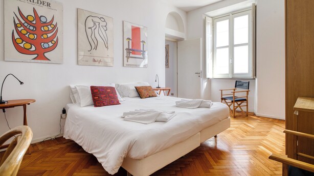 A bedroom with a large bed, white bedding, a wooden floor, and artwork on the walls. - San Vincenzo - Milano Centro Storico (Milano)