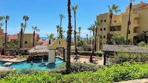 A resort pool area with palm trees, a thatched-roof structure, and a view of the ocean. - Brand New Listing: Elegant Studio Suite with Ocean and Pool View (Cabo San Lucas)