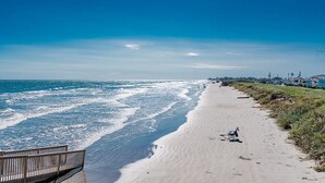Ein Strand mit einer Holzstege, der zum Wasser führt, ein klarer Himmel und einige Personen in der Ferne.