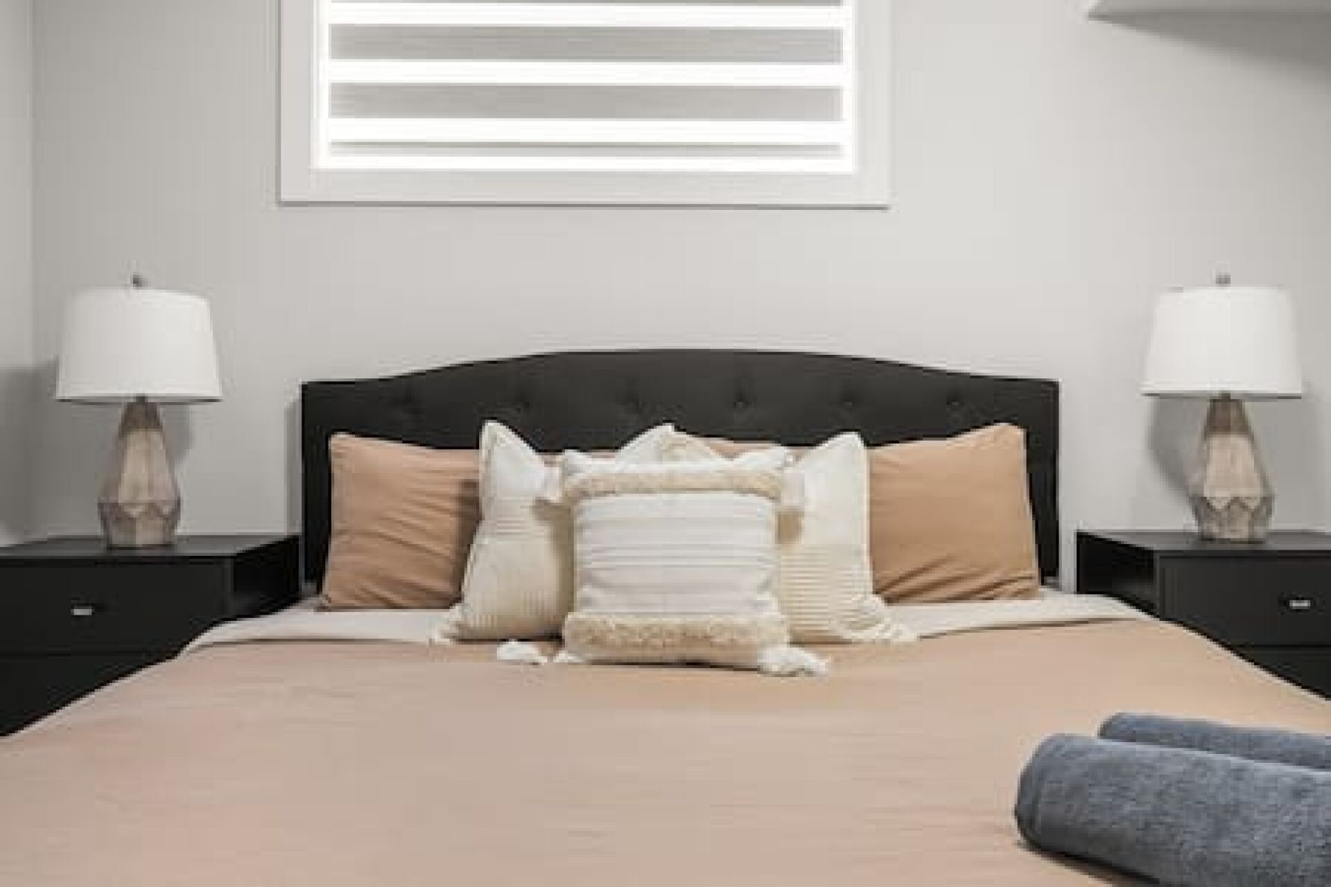 A neatly made bed with a black headboard, two bedside tables with lamps, and a window with blinds.