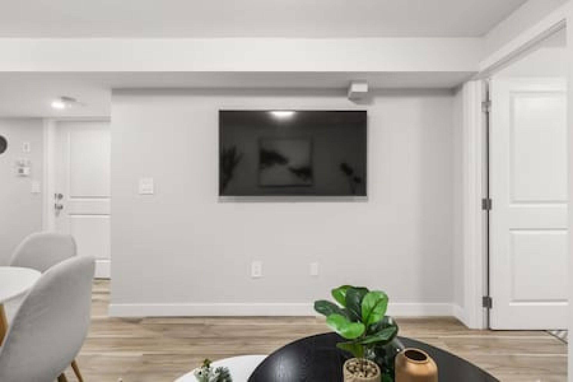A modern living room with a dining area, a large black artwork on the wall, and a potted plant on a black ottoman.