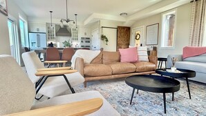 Un salon moderne avec un canapé en cuir marron, un fauteuil gris et une table basse noire. On y trouve également un coin repas avec une table et des chaises en bois, et une cuisine à l’arrière-plan.