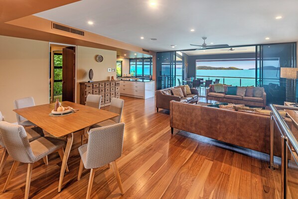 A modern living area with a wooden dining table, comfortable seating, a kitchen, and a balcony with a view of the sea. - ABSOLUTE WATERFRONT LUXURY WITH NEW BUGGY (Hamilton Island)