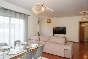 A modern living room with a glass dining table, a beige sofa, and a flat-screen TV mounted on the wall.