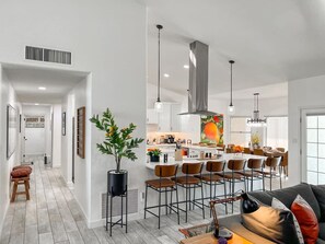 A modern kitchen with a central island, bar stools, pendant lighting, and a dining area with chairs.