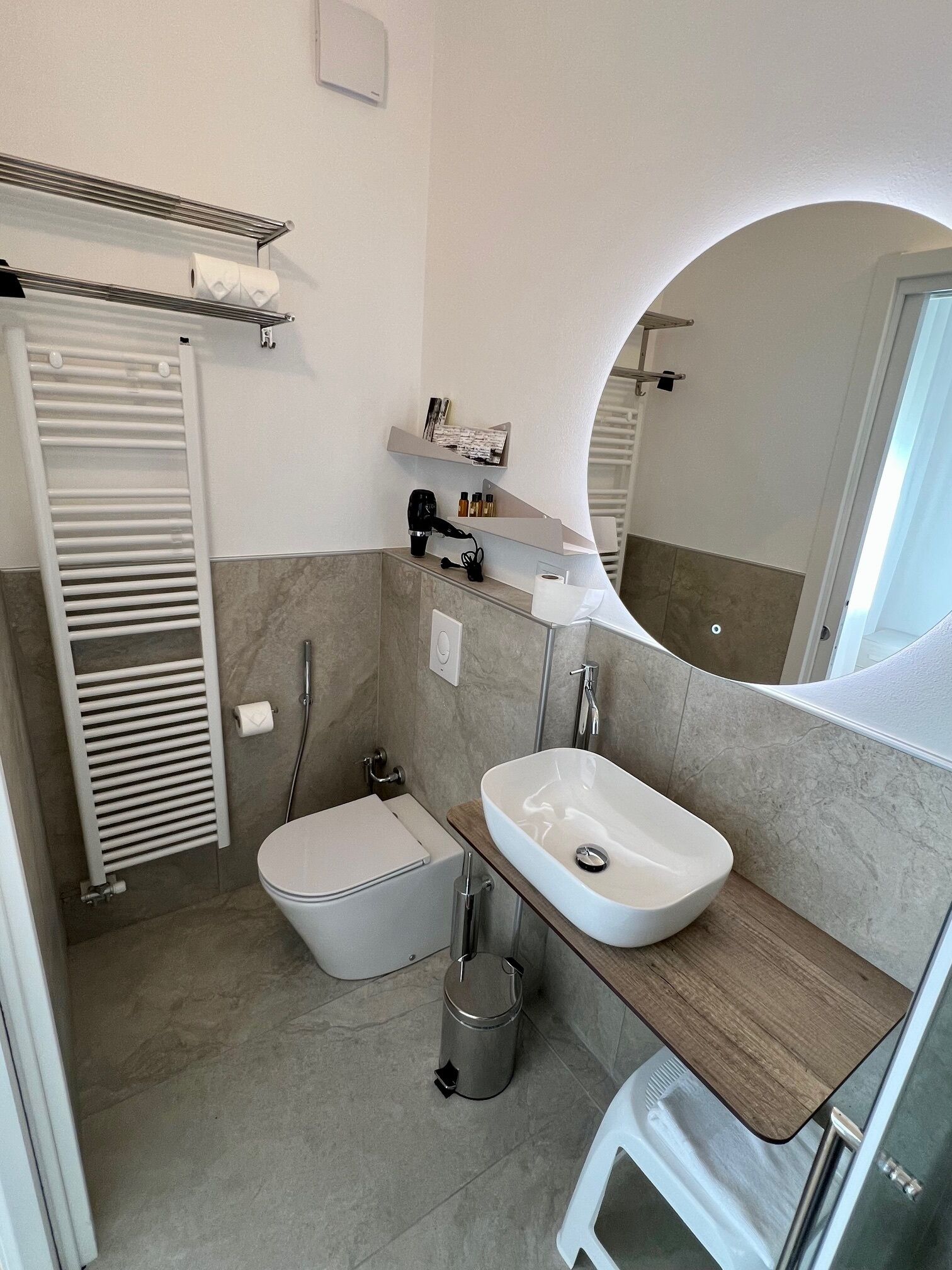Et moderne badeværelse med toilet, vask og brusebad. Der er et håndklædetørrestativ, et spejl og en lille hylde med toiletartikler.