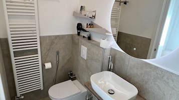 Et moderne badeværelse med toilet, vask og brusebad. Der er et håndklædetørrestativ, et spejl og en lille hylde med toiletartikler.