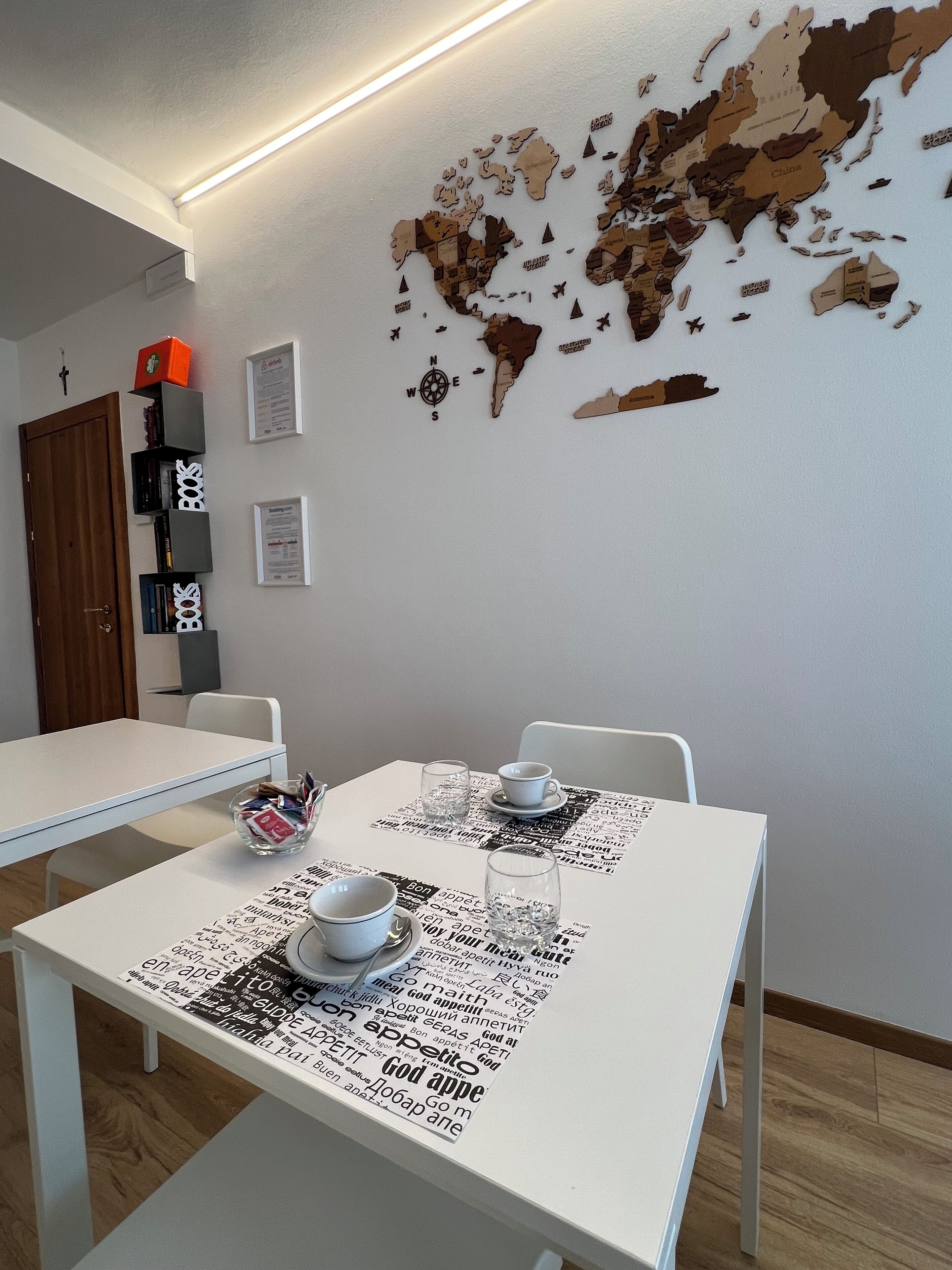 A dining area with a white table and chairs, a world map on the wall, and a bookshelf with books and a red box.