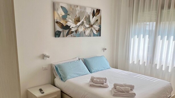 A modern bedroom with a bed, bedside tables, a nightstand, a colorful chair, and a large window with sheer curtains. - Ceciaìs House ROOM 2 (Bassano del Grappa)