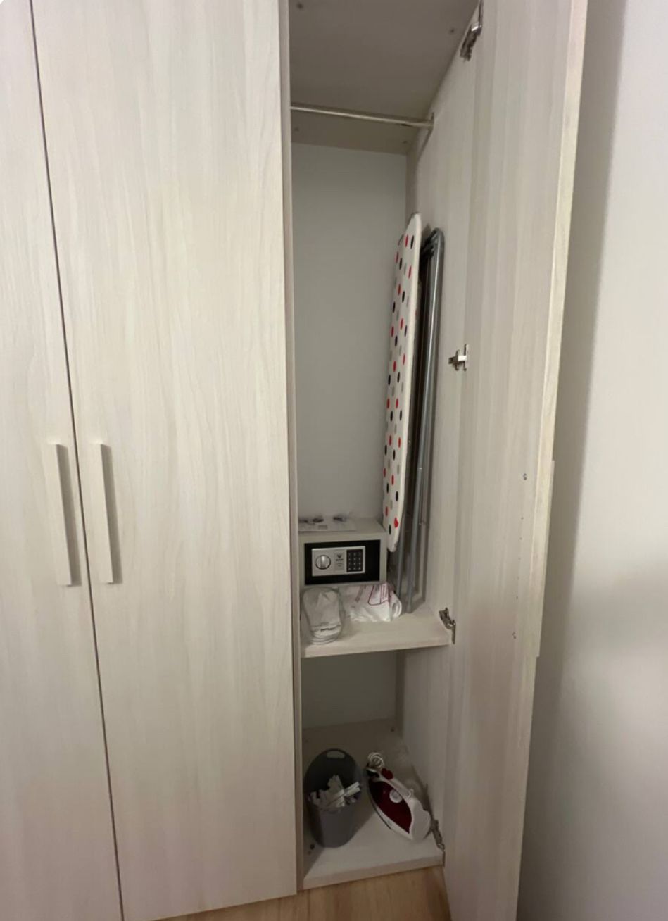 Ein Kleiderschrank mit Kleiderstange und Ablagefläche, inklusive Bügeleisen und kleiner Schachtel.