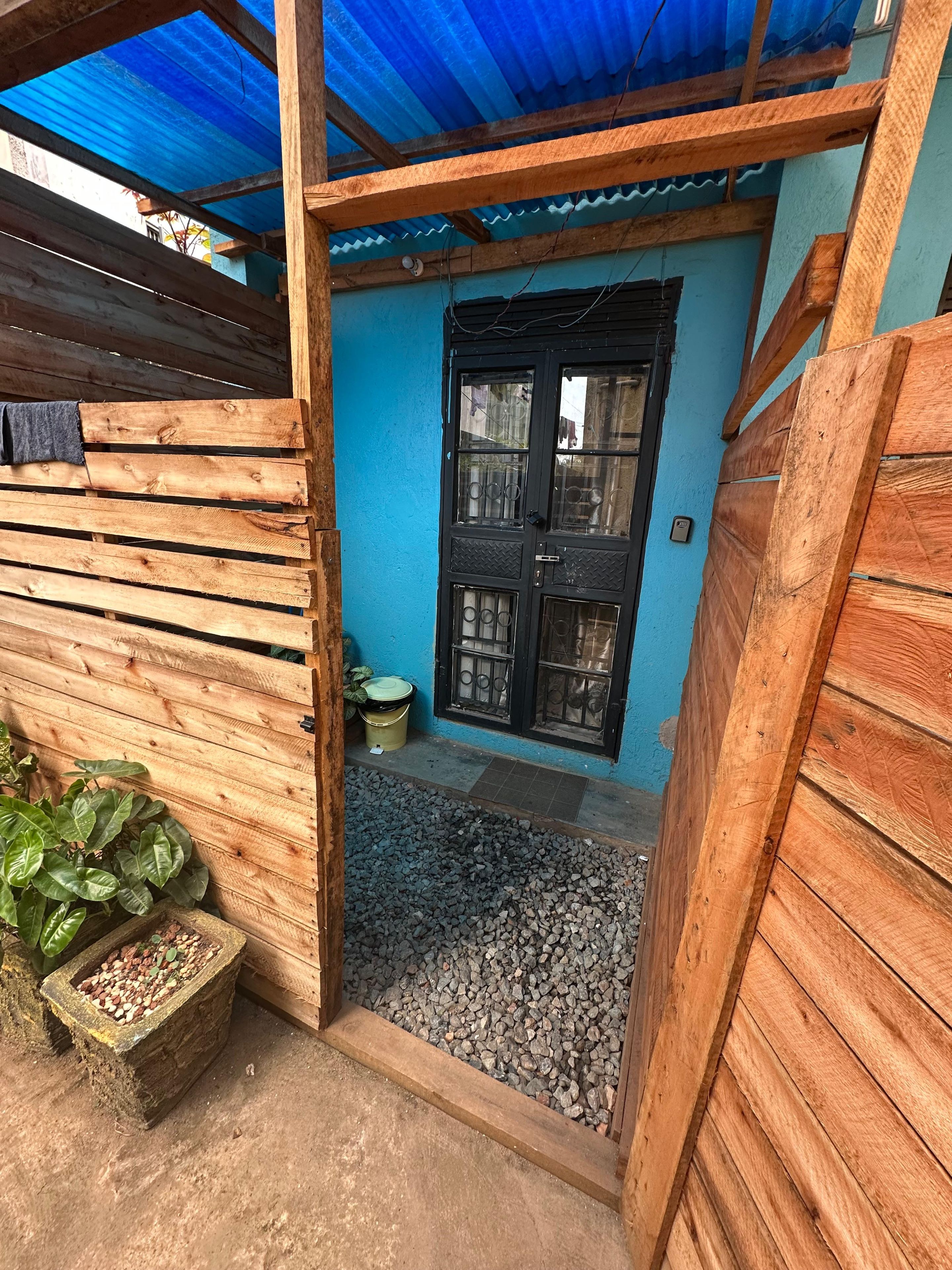 A wooden structure with a blue wall and a black door, a gravel path leading to the door, and a plant on the left.