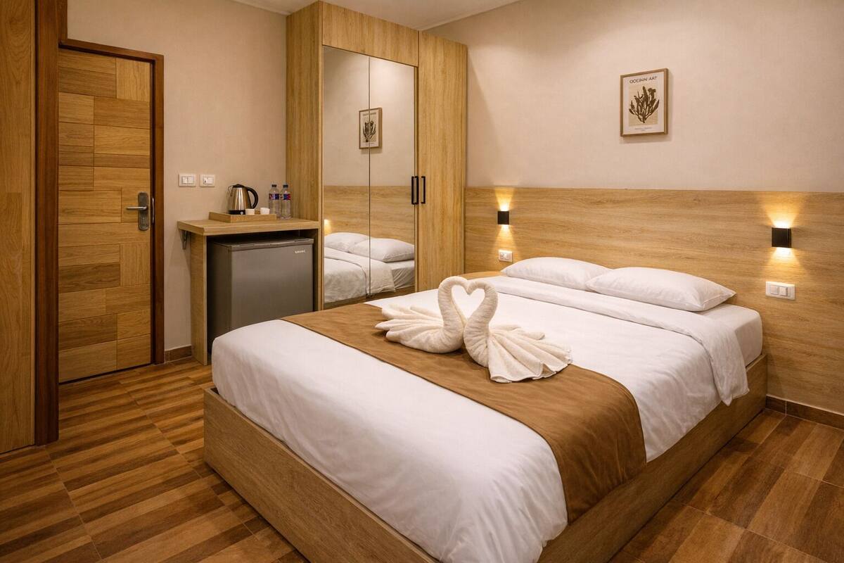 A hotel room with a wooden floor, a bed with white bedding, a minibar, a mirror, and two wall-mounted lights.