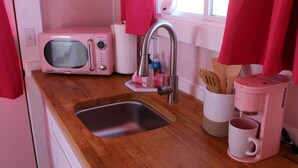 Cozinha com bancada de madeira, pia, micro-ondas rosa, cafeteira e janela com cortinas vermelhas.