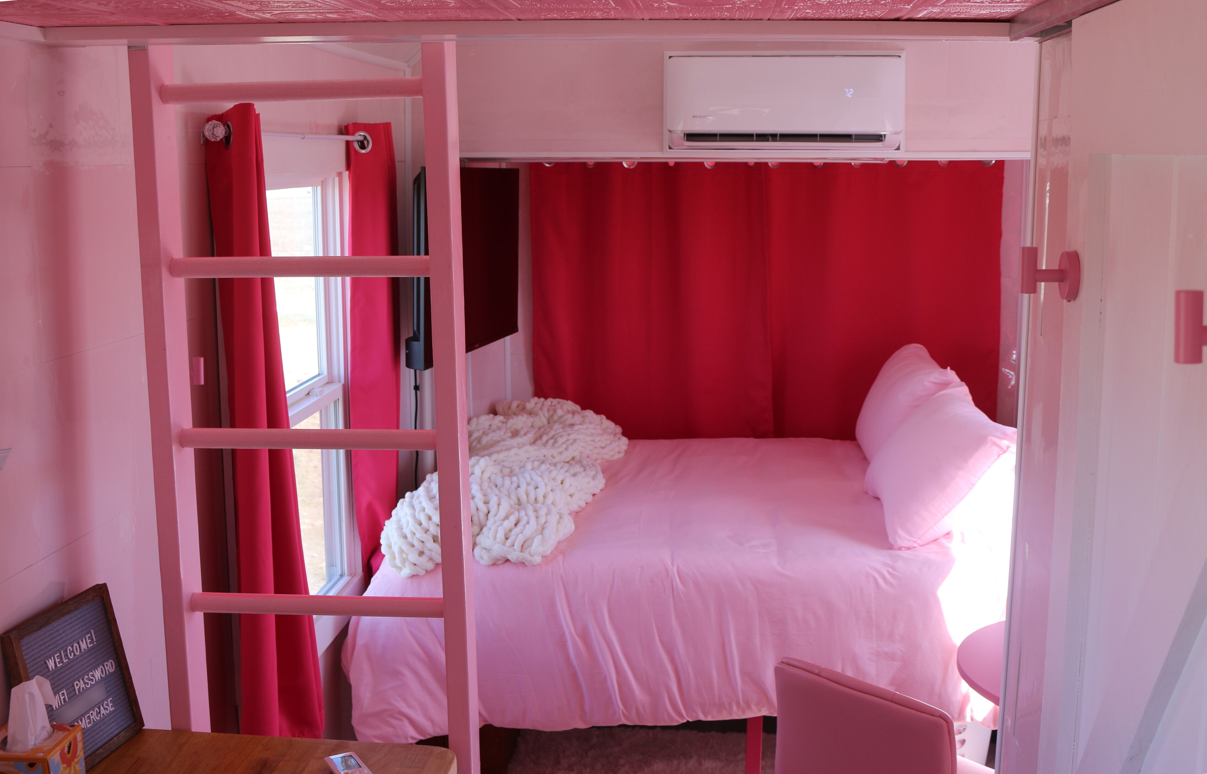 Een kamer in roze tinten met een stapelbed, een raam met rode gordijnen en een airconditioning.