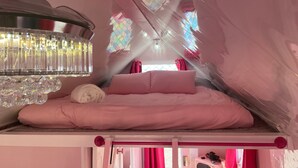 Quarto com decoração em tons de rosa, com uma cama beliche, um candelabro e uma janela com desenhos coloridos.