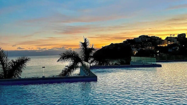 A pool with clear blue water, palm trees, and a sunset view of a coastal town. - Modern Studio with Ocean View (Cruz de Huanacaxtle)