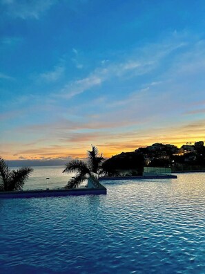 A pool with clear blue water, palm trees, and a sunset view of a coastal town. - Modern Studio with Ocean View (Cruz de Huanacaxtle)