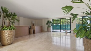 Um hall amplo com balcão de recepção, plantas em vasos grandes e vista para o exterior através de amplas janelas.
